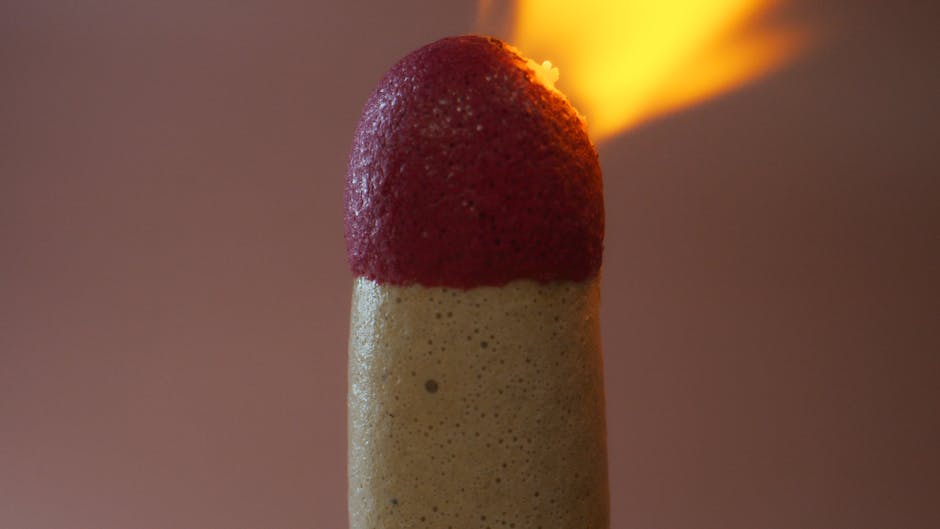 A tall, beige foam or sponge-like object with a rounded top, partially covered in a red, textured substance resembling berry or fruit topping. The object is positioned vertically on a flat surface inside a property, near the doorway, with a slightly blurred background. To the side, a portable flame torch with an orange-yellow flame is visible, indicating possible heat or combustion process. The scene appears to be indoors with soft lighting. This image may relate to packing or protective materials used during a home relocation, as seen in the context of removals services offered by Man and Van Leyton, focusing on furniture transport and moving logistics.