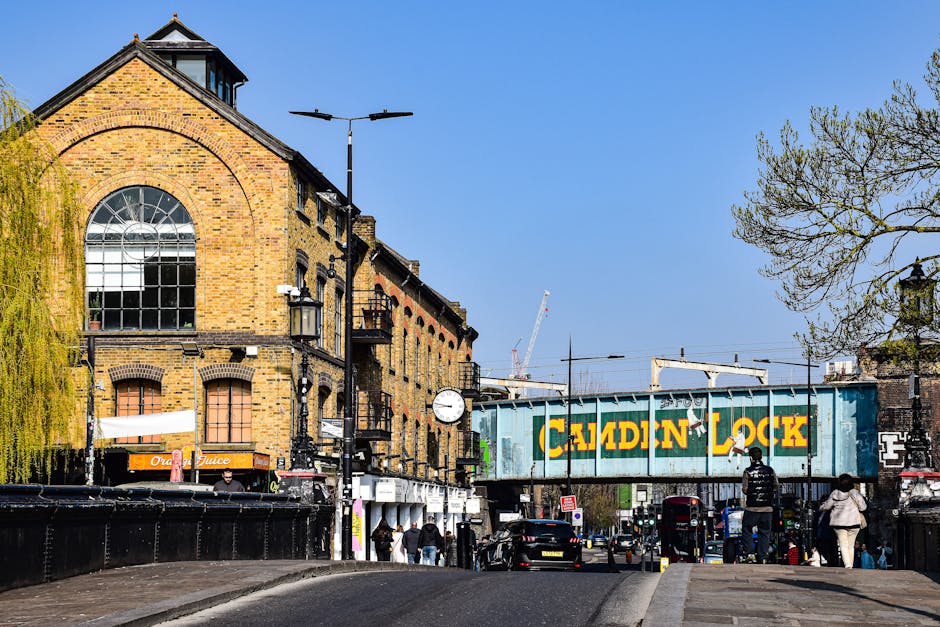 A street view in Leyton showing a brick building with large arched windows and a clock attached to its facade. In the foreground, several pedestrians walk along the pavement, some near a black fence o