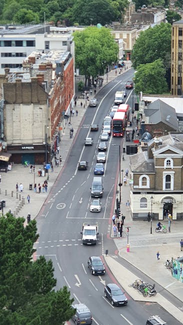 A street view in Leyton showing a brick building with large arched windows and a clock attached to its facade. In the foreground, several pedestrians walk along the pavement, some near a black fence o