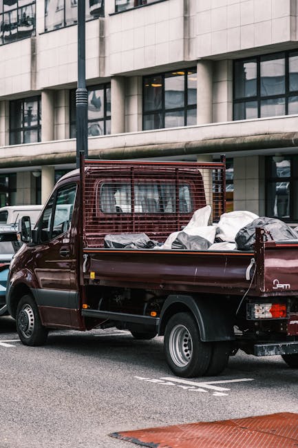 A burgundy flatbed utility vehicle parked on a city street in Leyton, featuring a metal mesh side panel and filled with various household items wrapped in plastic and fabric, including boxes and bags. The vehicle is positioned next to a modern building with large glass windows and a concrete facade, with a black lamp post nearby and white vehicles parked in the background. The scene depicts the loading or unloading stage of a home relocation or furniture transport process, with the vehicle ready for moving delivery services by [COMPANY_NAME]. The street has clear markings, and the overall environment suggests a busy urban area suitable for professional removals or house moving activities in Leyton, LEYTON MIDLAND ROAD, as detailed in the removals pickup guide.