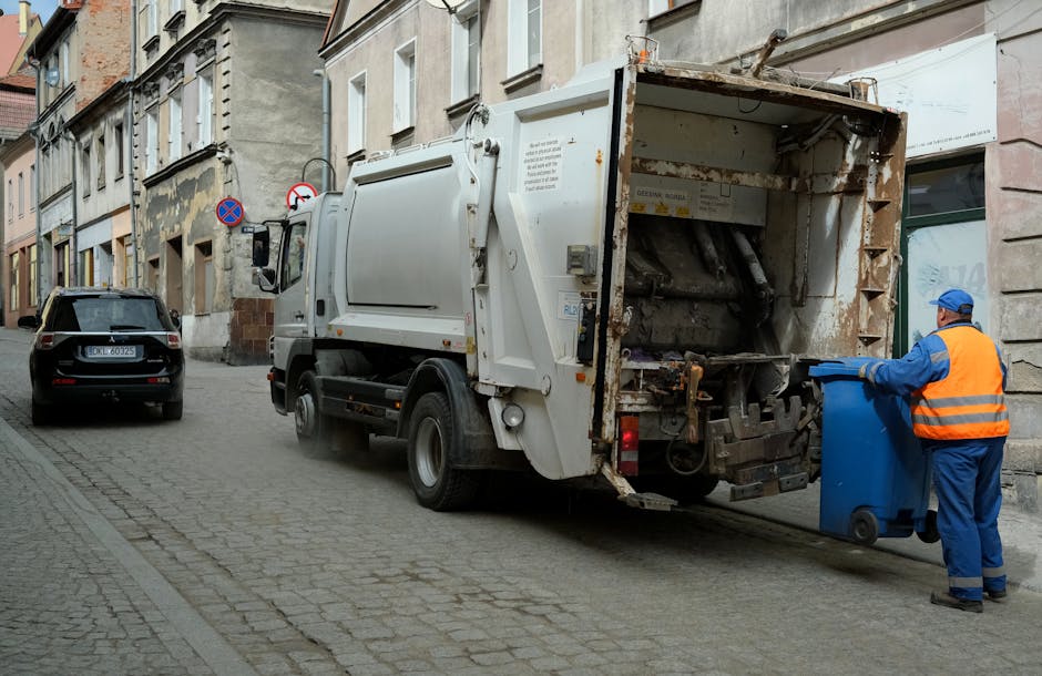 A waste collection truck parked on a narrow cobbled street outside a residential building, with an employee in a high-visibility orange vest and blue uniform operating a blue wheeled bin, about to load it into the rear compartment of the garbage truck. The truck's rear is open, revealing empty mechanical compartments used for compacting waste. Nearby, there is a black car parked facing away from the truck, along with urban buildings lining the street, some with peeling paint and old brickwork. The scene takes place during daylight, with natural lighting illuminating the surroundings, and the area appears to be part of a local urban resi­dent­ial district, relevant to home relocation or waste collection services included in [SERVICE_CATEGORY], as highlighted in the Leyton Midland Road removals pickup guide from [PAGE_TITLE] on [PAGE_URL], with [COMPANY_NAME] involved in waste transport and logistics.